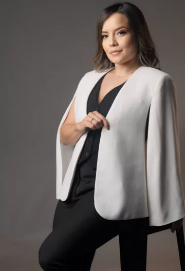A imagem mostra uma mulher de pele clara e cabelos castanhos, lisos e na altura dos ombros. Ela está vestindo uma roupa elegante, composta por um blazer branco com um design diferenciado, que parece uma capa, e uma blusa preta por baixo. Ela também está usando calças pretas. A mulher está em pé, com uma mão apoiada no quadril e a outra segurando a lapela do blazer. O fundo da imagem é neutro, em tons de cinza.